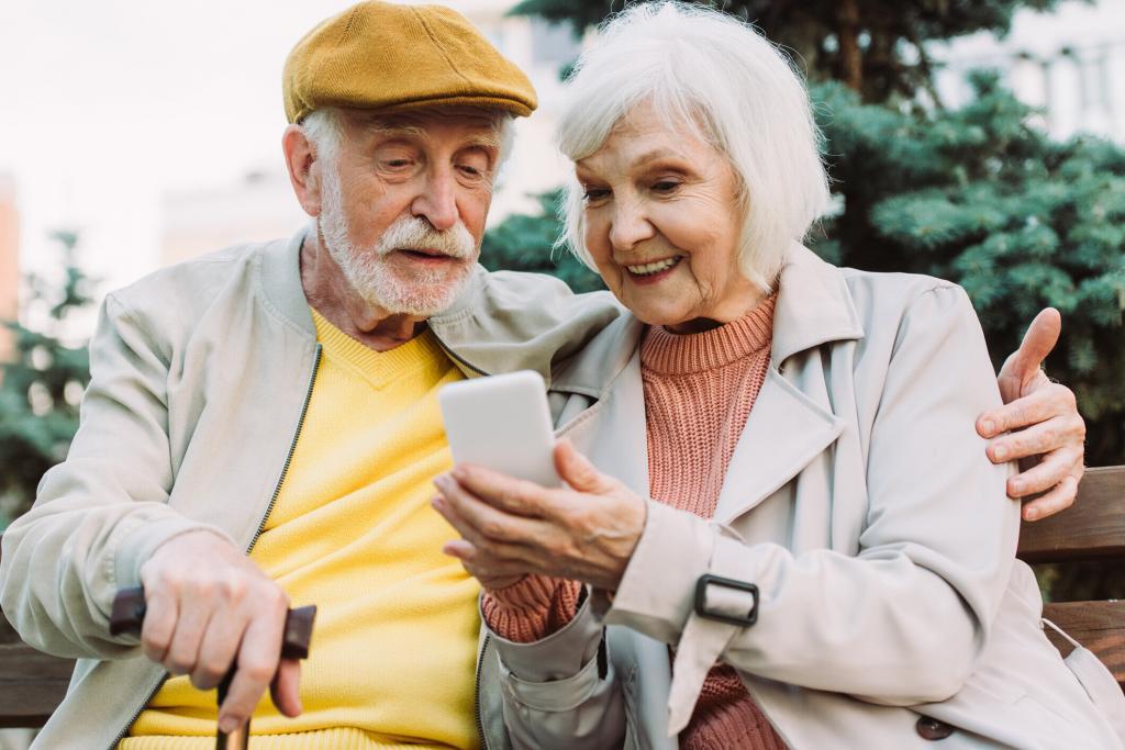 Selective focus of smiling senior woman holding smartphone near husband on bench in park Senior Couple