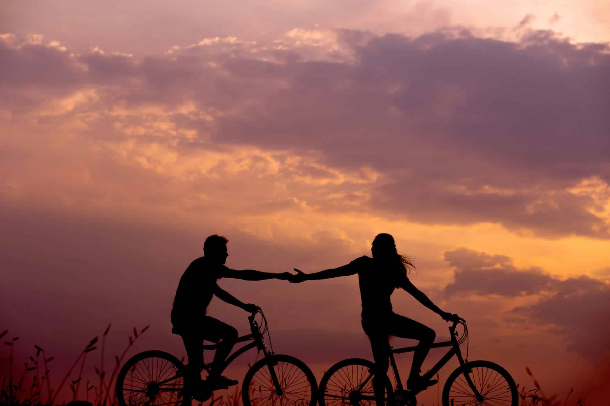 Two people on bikes touching hands in front of a cloudy sunset
