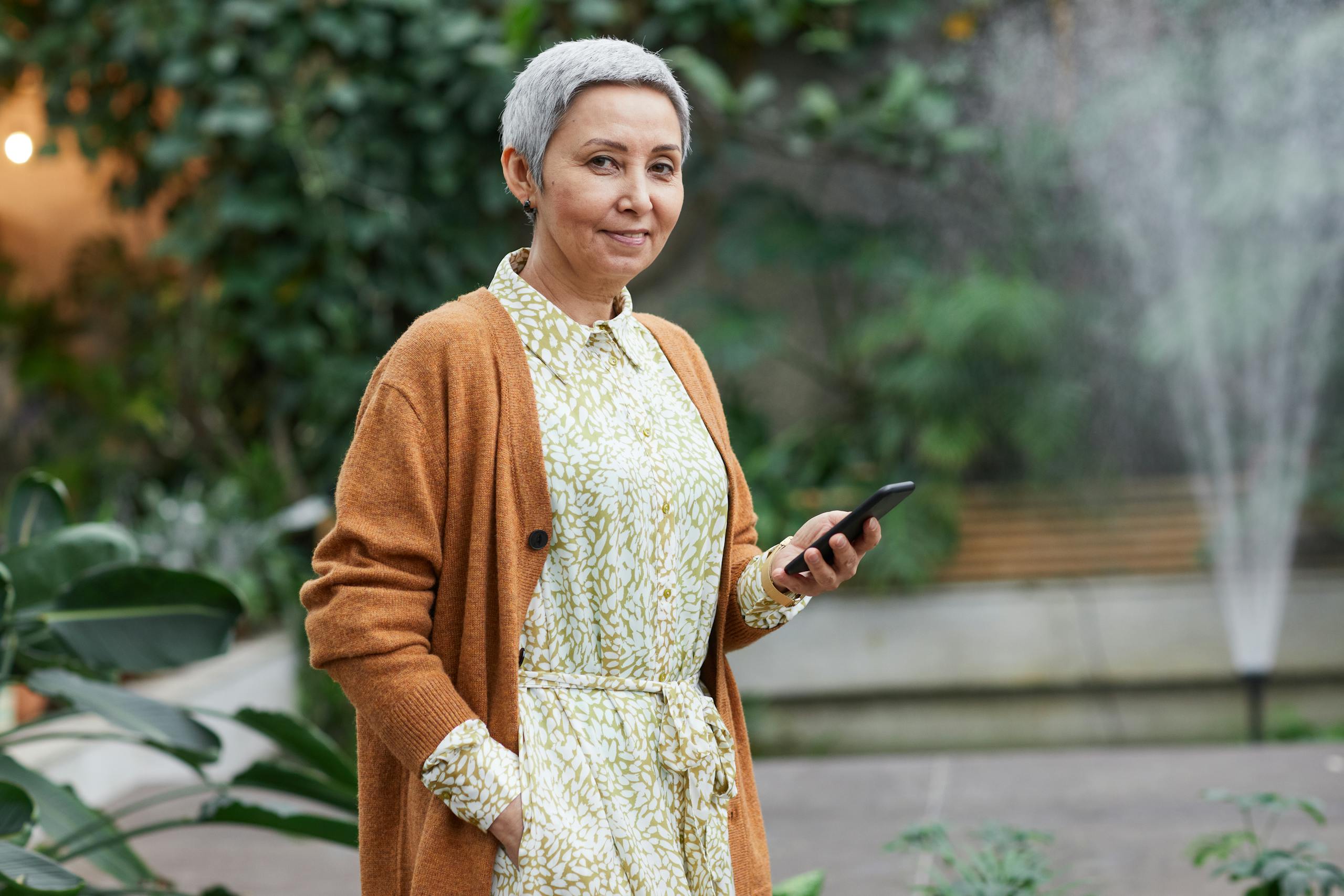 Senior woman smiling while using a smartphone in a garden setting, with greenery in the background.